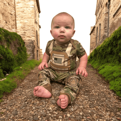 Baby in Multicam Tactical Uniform with flak jacket print and US flag, sitting on stone path surrounded by greenery.