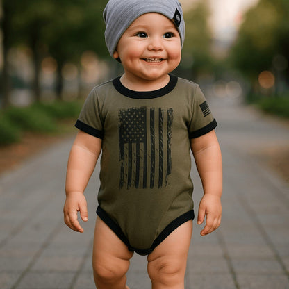 Patriotic baby bodysuit in OD Green with distressed American flag design, worn by a smiling toddler outdoors.